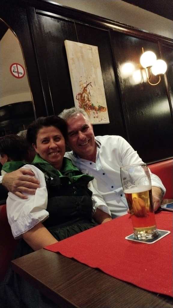 Eine Frau in traditioneller Kleidung und ein Mann sitzen lächelnd in einem Restaurant, vor ihnen steht ein Glas Bier auf dem Tisch.