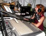 Eine Frau sitzt an einem Mischpult in einem Radio-Studio und trägt Kopfhörer, während sie spricht.